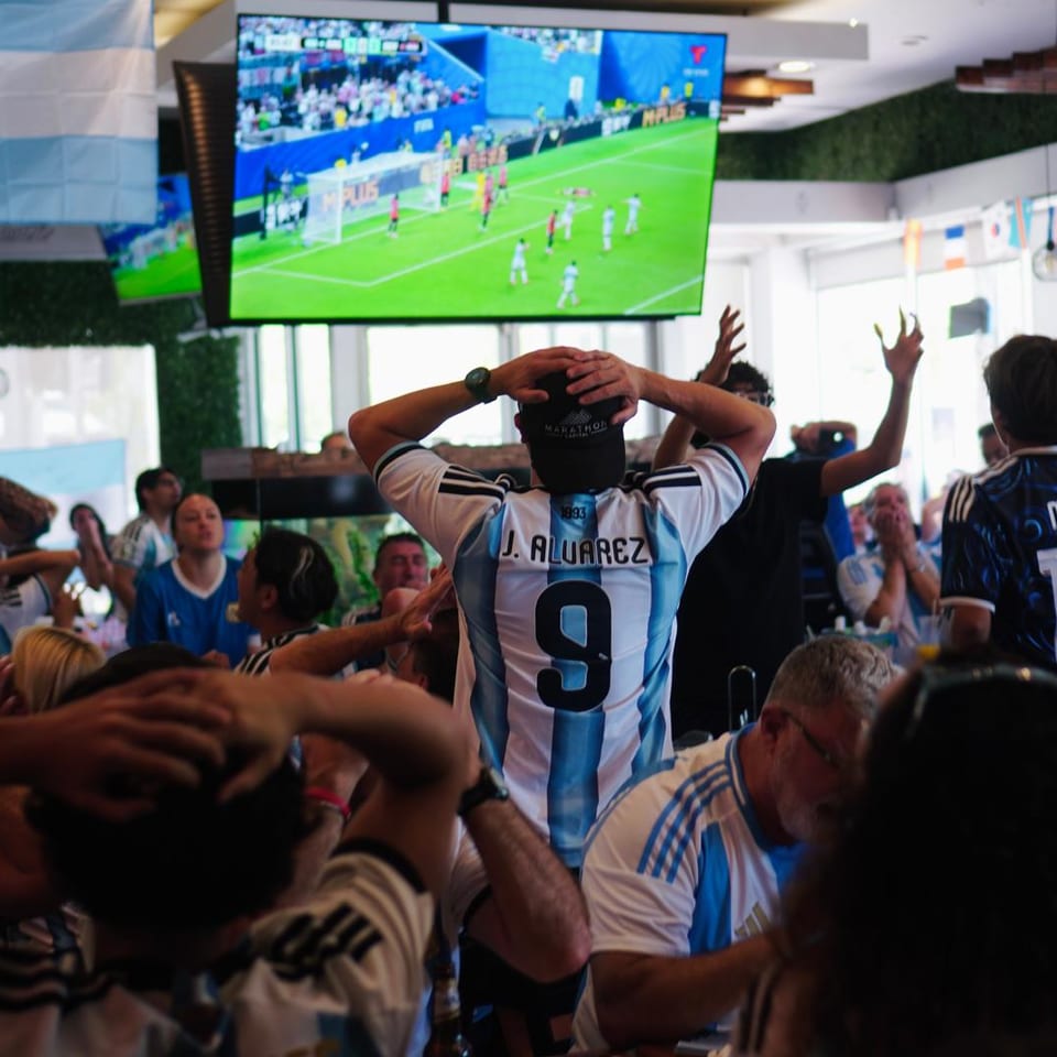 Fans in Argentina jerseys reacting to a televised fútbol match inside Manolo.