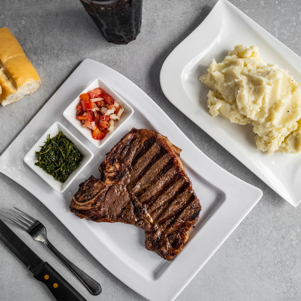 Grilled steak served with mashed potatoes, sautéed greens, diced tomatoes, and sliced bread on white plates.