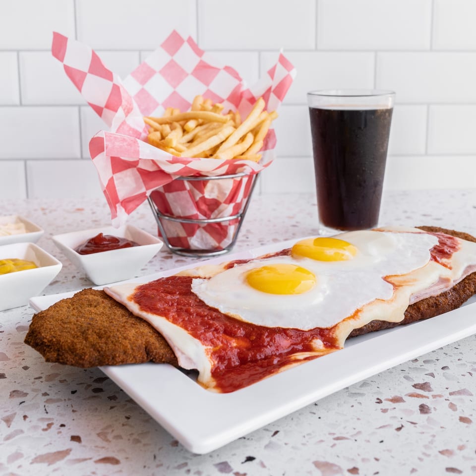 Breaded steak topped with tomato sauce and fried eggs, served with fries, dipping sauces, and a glass of soda.